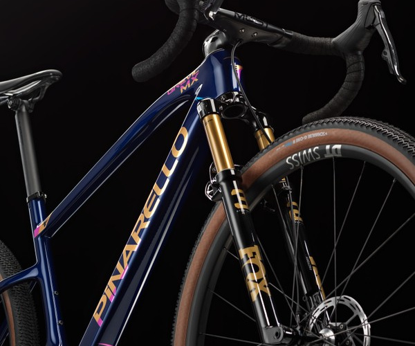 A close-up view of a Pinarello bicycle featuring a sleek navy blue frame with gold and pink accents. The bike has a prominent front fork with a suspension system and disc brakes, as well as textured tires designed for various terrains. The image is set against a dark background, highlighting the bike's design and details.