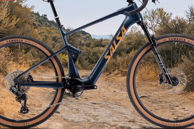 Image of a Salsa bike model, titled "Wanderosa," positioned on a rugged, natural trail surrounded by greenery. The bike features a sleek blue frame with distinctive orange branding. The foreground showcases the bike's detailed components, emphasizing its design suited for off-road adventures. The sky above is partly cloudy, enhancing the outdoor atmosphere.