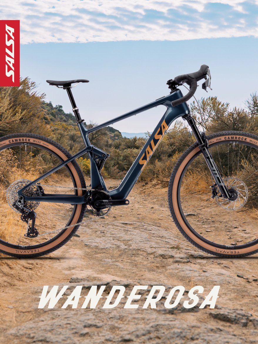 Image of a Salsa bike model, titled "Wanderosa," positioned on a rugged, natural trail surrounded by greenery. The bike features a sleek blue frame with distinctive orange branding. The foreground showcases the bike's detailed components, emphasizing its design suited for off-road adventures. The sky above is partly cloudy, enhancing the outdoor atmosphere.