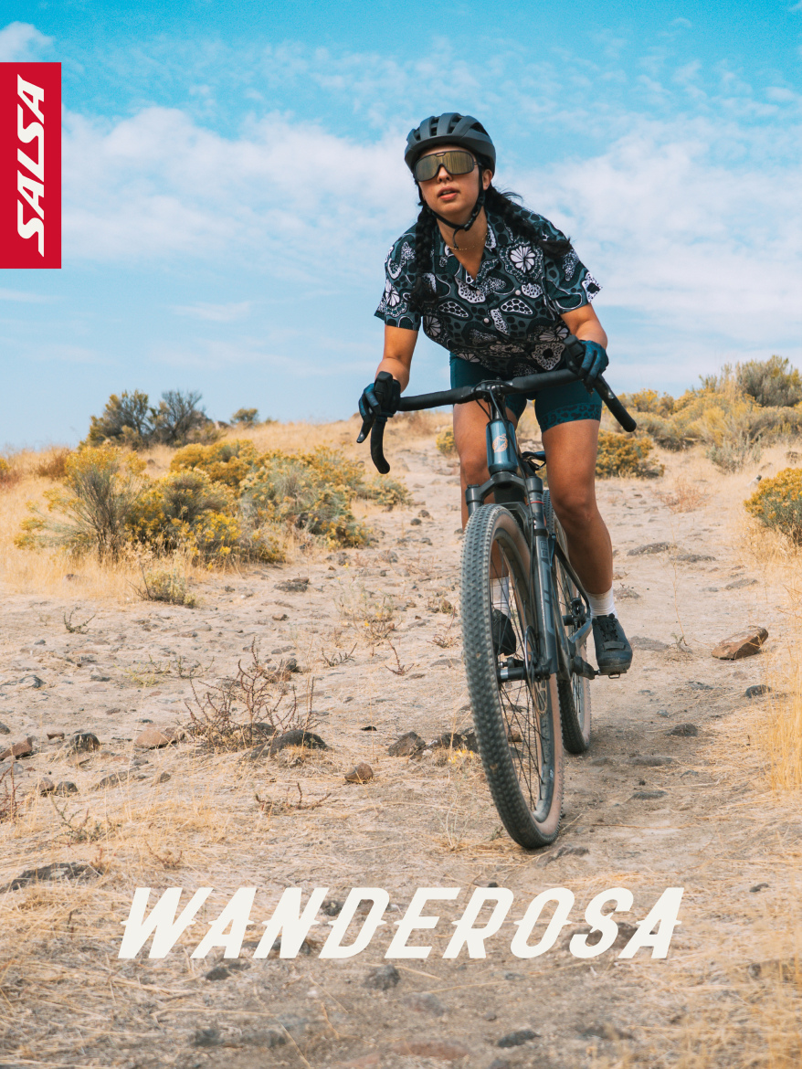 A person riding a gravel bike on a rugged trail surrounded by dry shrubs and grass. They are wearing a patterned cycling shirt, shorts, a helmet, and sunglasses, focused on navigating the terrain. The sky is bright with a few clouds, and there is a logo in the top left corner. The text "WANDEROSA" is prominently displayed at the bottom of the image.