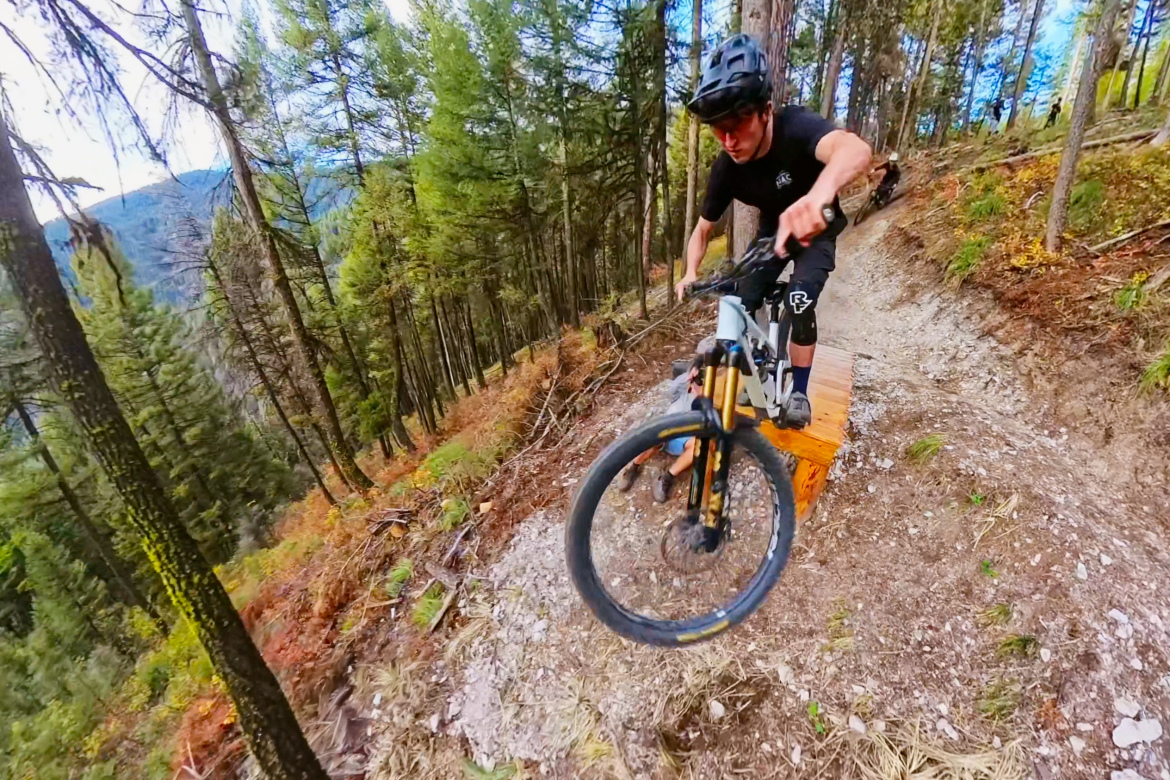 A mountain biker wearing a black helmet and shirt is performing a jump over a wooden ramp on a trail surrounded by tall trees. The bike's front wheel is elevated above the ground, showcasing the cyclist's skill and focus while navigating the rugged terrain. In the background, additional trees and a mountainous landscape are visible, highlighting the outdoor setting.
