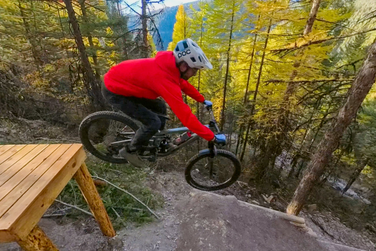A mountain biker in a red hoodie can be seen airborne over a dirt jump, with trees in vibrant fall colors in the background. The jump is made of dirt and is located in a forested area, showcasing an outdoor adventure setting.