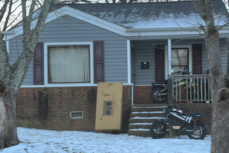 A gray house with maroon shutters and a wooden porch, featuring a set of stairs leading up to the front door. In front of the house, there is a yellow refrigerator positioned beside the stairs, and two motorcycles are parked on the porch. The ground is covered with a light layer of snow, and there are trees in the foreground.