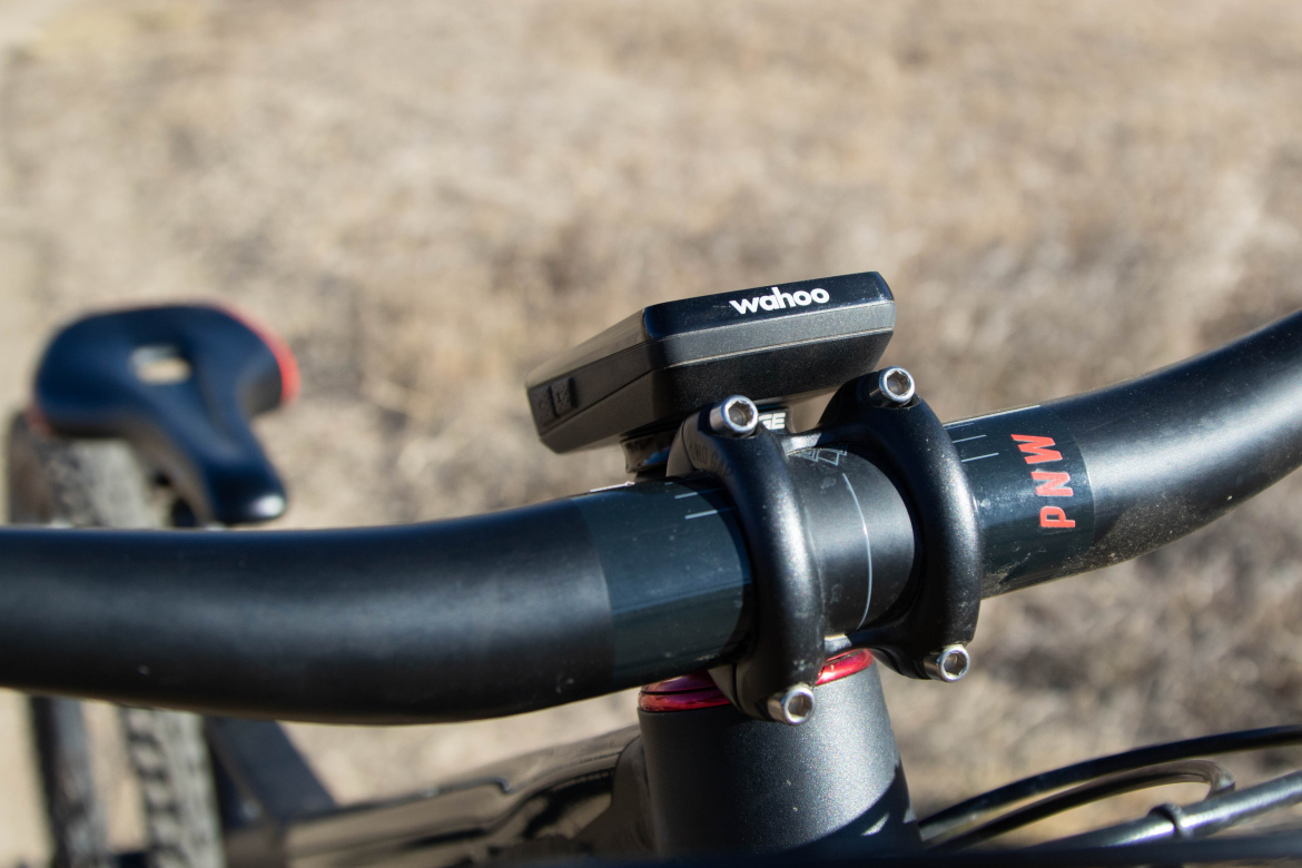 Close-up view of a bicycle handlebar featuring a Wahoo cycling computer mounted on the stem, with a blurred background of dry grassland. The handlebar is black, with a red accent on the stem, highlighting the PNW brand.