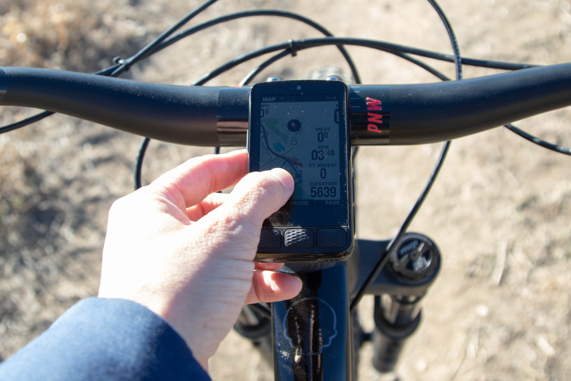 A close-up view of a hand adjusting a device mounted on a bicycle handlebar, displaying a GPS screen with ride statistics and a map, against a background of dirt and grass.