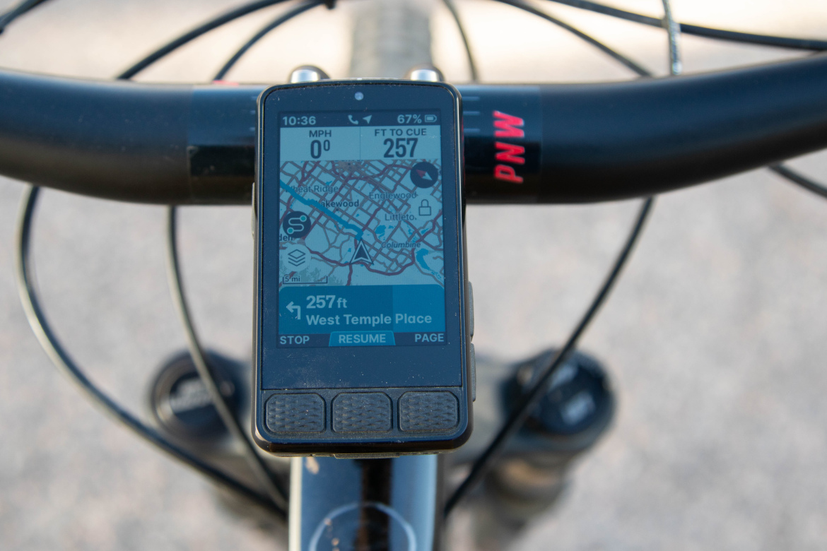 Close-up view of a bicycle handlebar with a GPS device mounted on it, displaying a navigation map. The screen shows the current speed of 0 mph, elevation of 257 feet, and directions to "West Temple Place." The time reads 10:36, and the battery level is at 67%.