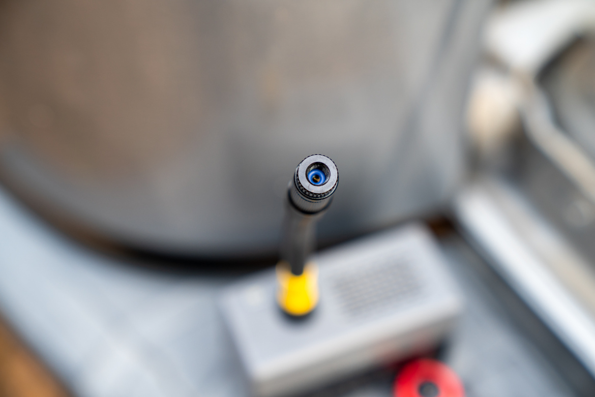 Close-up of the nozzle of a pressure washer, displaying a black tip with a blue center, set against a blurred background of cleaning equipment.