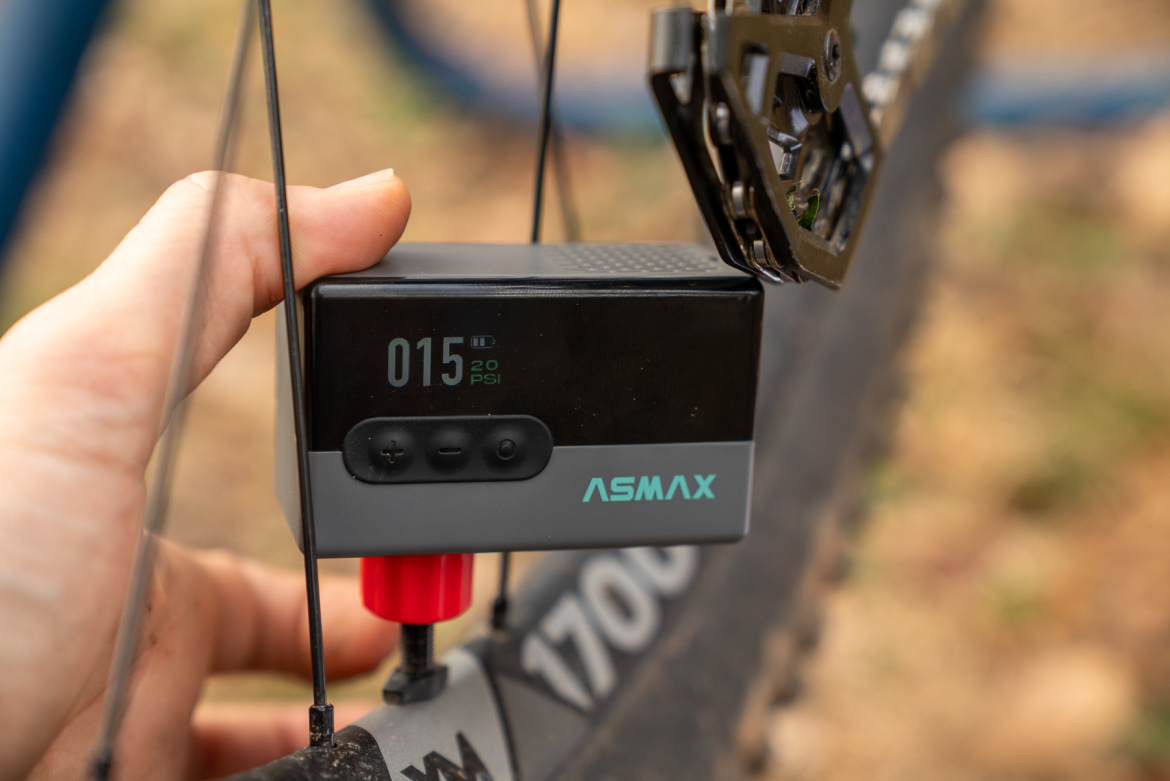 A close-up image of a hand holding a digital tire pressure gauge. The display shows a reading of 15 PSI and a battery indicator, with the ASMAX logo visible on the device. The gauge is connected to a bike tire, with part of the bike frame and tire tread visible in the background.