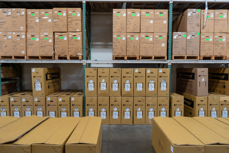 A warehouse setting with multiple stacked cardboard boxes on metal shelves, labeled with various green and yellow stickers. The lower section features additional boxes stacked on the ground, all with labels indicating their contents. The boxes appear to be organized for inventory or shipping purposes, with handwriting visible on some boxes.