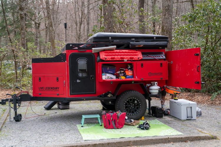 A vibrant red camping trailer parked in a wooded area, with one side open showcasing camping gear and supplies. A green mat is laid out on the ground, with backpacks and other items scattered around, and a small step stool nearby. The trailer is equipped with off-road wheels and a top-mounted storage box. Trees and shrubs surround the scene, creating a natural camping environment.
