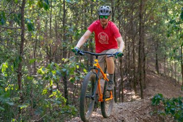 A person riding a mountain bike on a gravel trail surrounded by trees. The rider wears a red shirt, helmet, and sunglasses, navigating through a wooded area with greenery all around. Craighead Forest Park mountain bike trail.