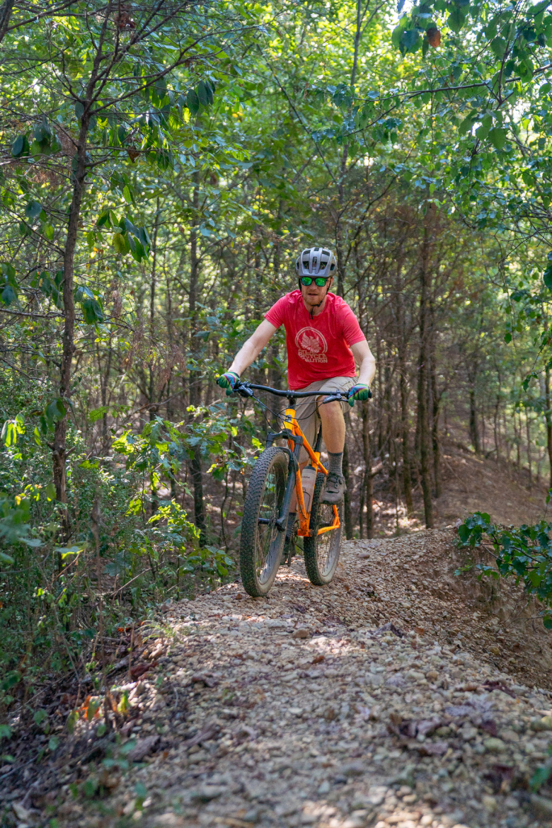 A person riding a mountain bike on a gravel trail surrounded by trees. The rider wears a red shirt, helmet, and sunglasses, navigating through a wooded area with greenery all around. Craighead Forest Park mountain bike trail.