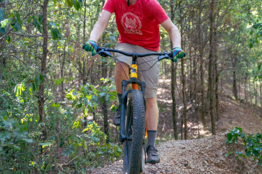 A person riding a mountain bike on a narrow gravel trail surrounded by trees, wearing a red t-shirt, sunglasses, and a helmet. The cyclist is focused, navigating through the wooded area on a sunny day. Craighead Forest Park mountain bike trail.