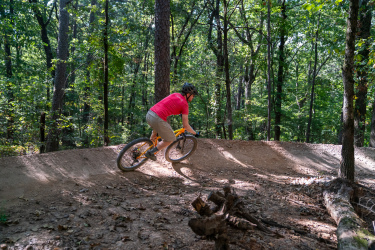 A mountain biker in a red shirt and beige shorts navigates a dirt trail surrounded by trees in a wooded area, demonstrating an action-packed moment of the sport. Craighead Forest Park mountain bike trail.