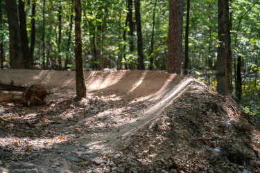 A winding dirt trail in a wooded area, featuring a smooth, banked curve and surrounded by trees. Sunlight filters through the foliage, casting gentle shadows on the path. Small rocks and leaves are scattered along the ground, enhancing the natural forest scenery. Craighead Forest Park mountain bike trail.