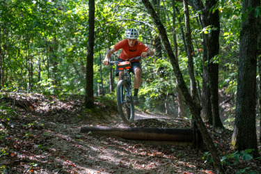 A mountain biker wearing a helmet and an orange shirt jumps over a wooden log on a forest trail, surrounded by lush green trees and foliage. Craighead Forest Park mountain bike trail.
