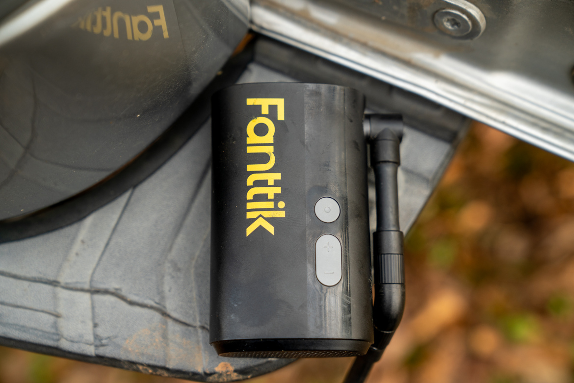 A close-up view of a portable device with the brand name "Fanttik" in bold yellow letters. The device features buttons for power and volume on the side and a connector attached to it. The device is placed on a textured surface with hints of autumn foliage in the background.