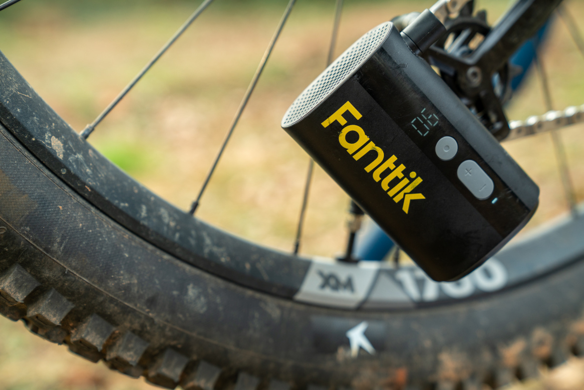 A close-up image of a Fanttik portable speaker attached to a bicycle tire, showcasing the speaker's controls and display against a blurred outdoor background. The bicycle's tread and rim are partially visible, indicating an outdoor setting.