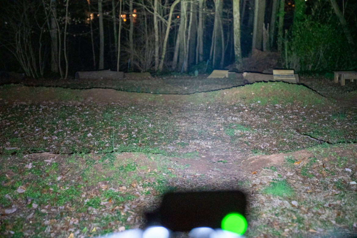 A nighttime outdoor scene showing a grassy area with scattered leaves and slight mounds. In the background, there are wooden ramps, and the edges of a forest can be seen with trees partially illuminated. The foreground features a blurry object with a green light, possibly a camera or flashlight.