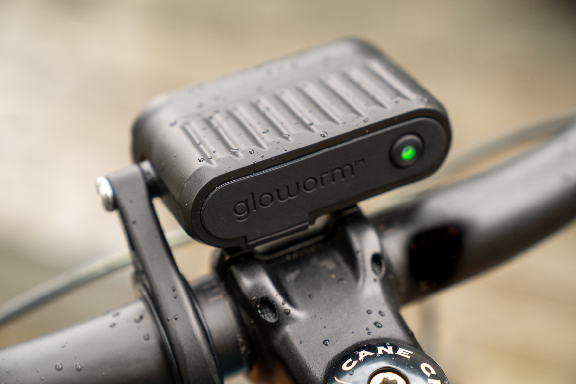 A close-up image of a black Glowworm bike light mounted on a bicycle handlebar. The surface of the light is wet, indicating it may be raining. The handlebar features a visible Cane Creek branding.