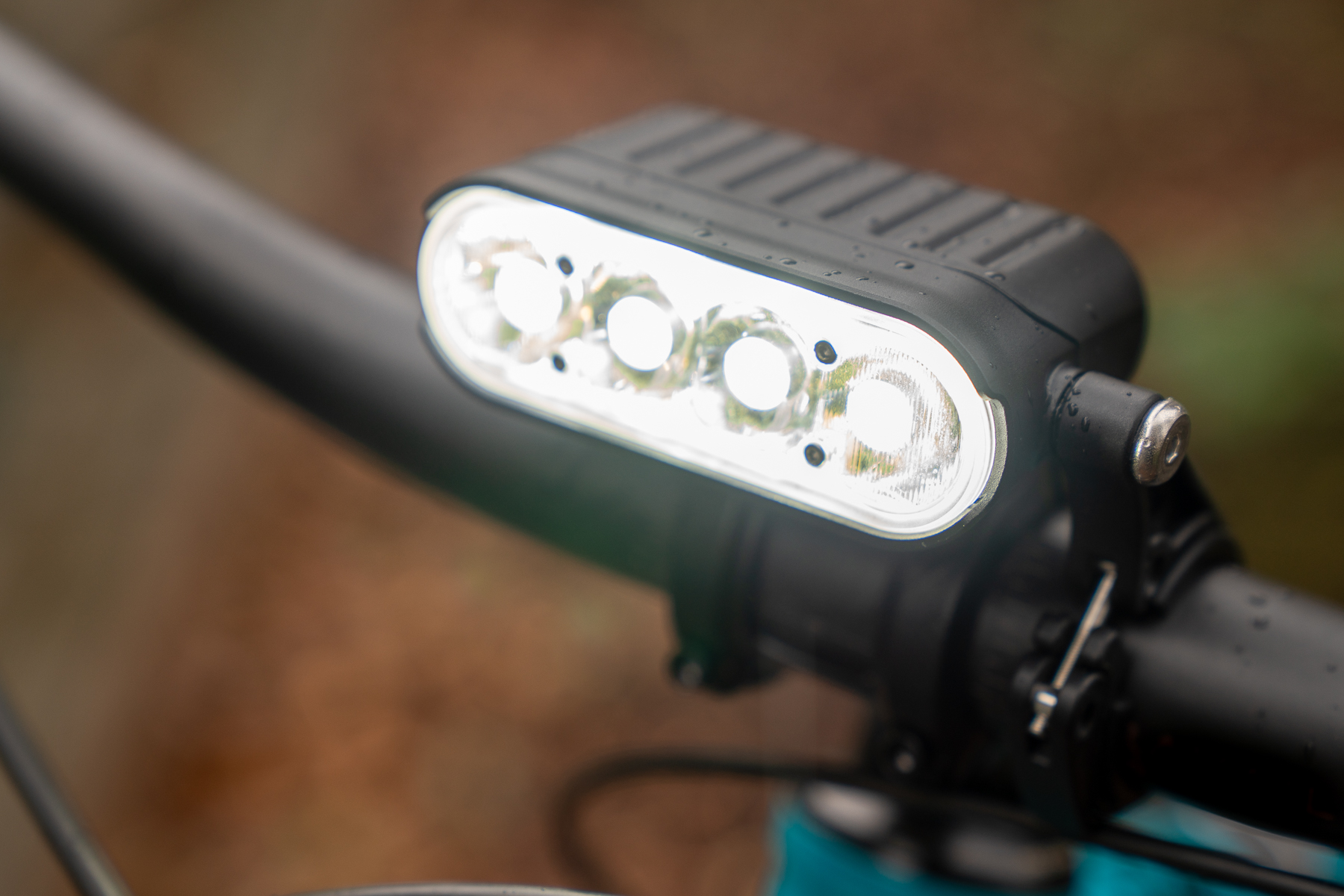 Close-up of a bicycle handlebar with a mounted front light featuring three LED bulbs, designed for visibility and safety. The light is waterproof, with droplets visible on its surface, and is attached securely to the handlebar. The background shows a blurred natural setting.