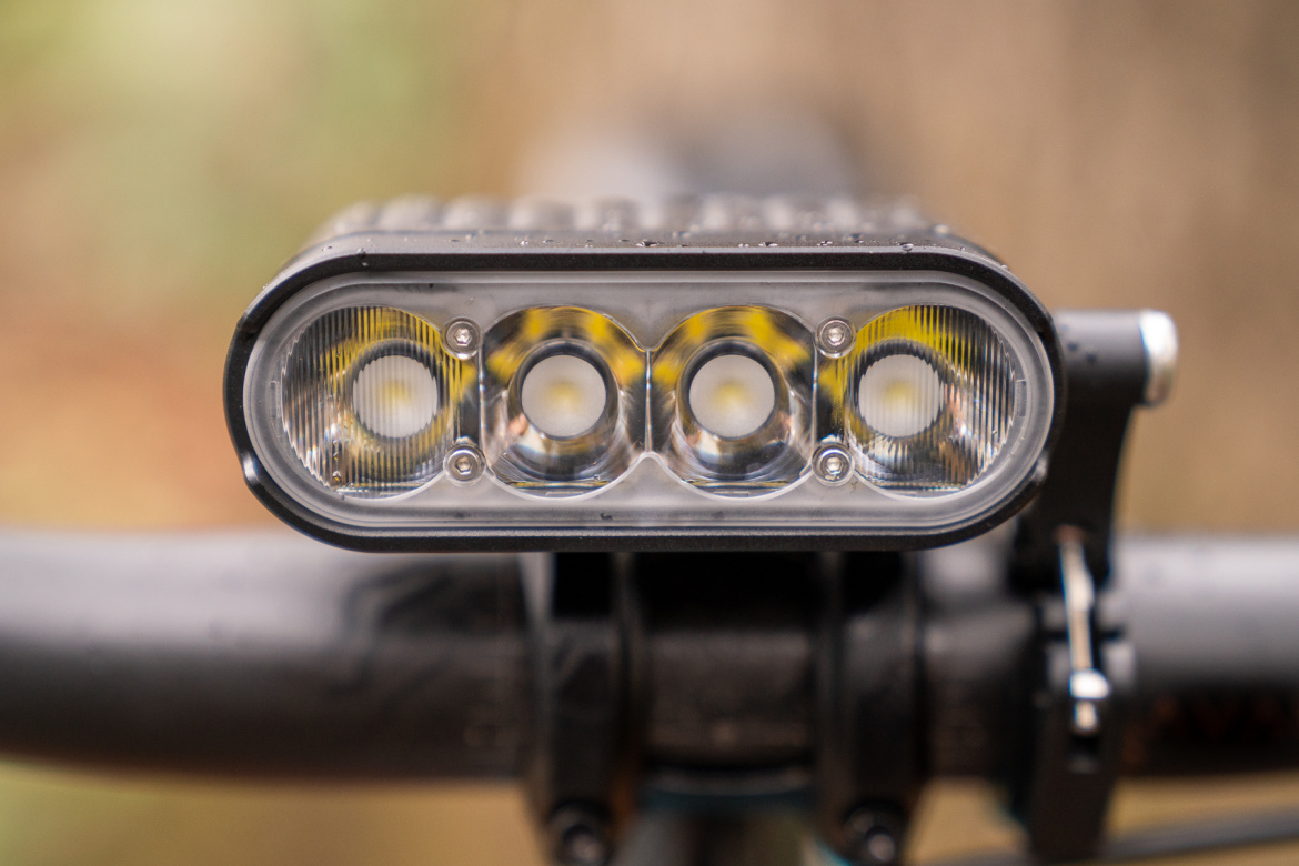 A close-up view of a bike front light mounted on the handlebars, featuring three bright LED lights within a sleek, waterproof casing. The background is blurred, suggesting an outdoor environment.