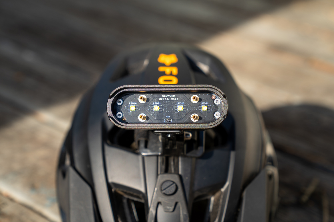 Close-up view of a bicycle helmet light attachment, featuring a sleek, rounded design with multiple LED indicators labeled LED10, LED11, LED20, and LED21. The light is mounted on top of a black helmet, with the brand name "Glowworm" visible. The background shows a rustic wooden surface.