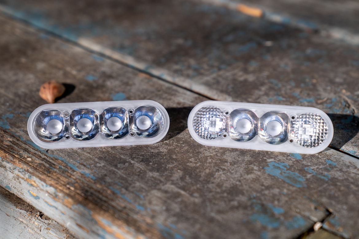 Two rectangular LED light units are positioned on a weathered wooden surface. Each unit features three circular lens openings arranged in a row, with one unit displaying a clear lens design and the other a textured lens design. A small nut sits nearby on the surface, highlighting the rustic setting.