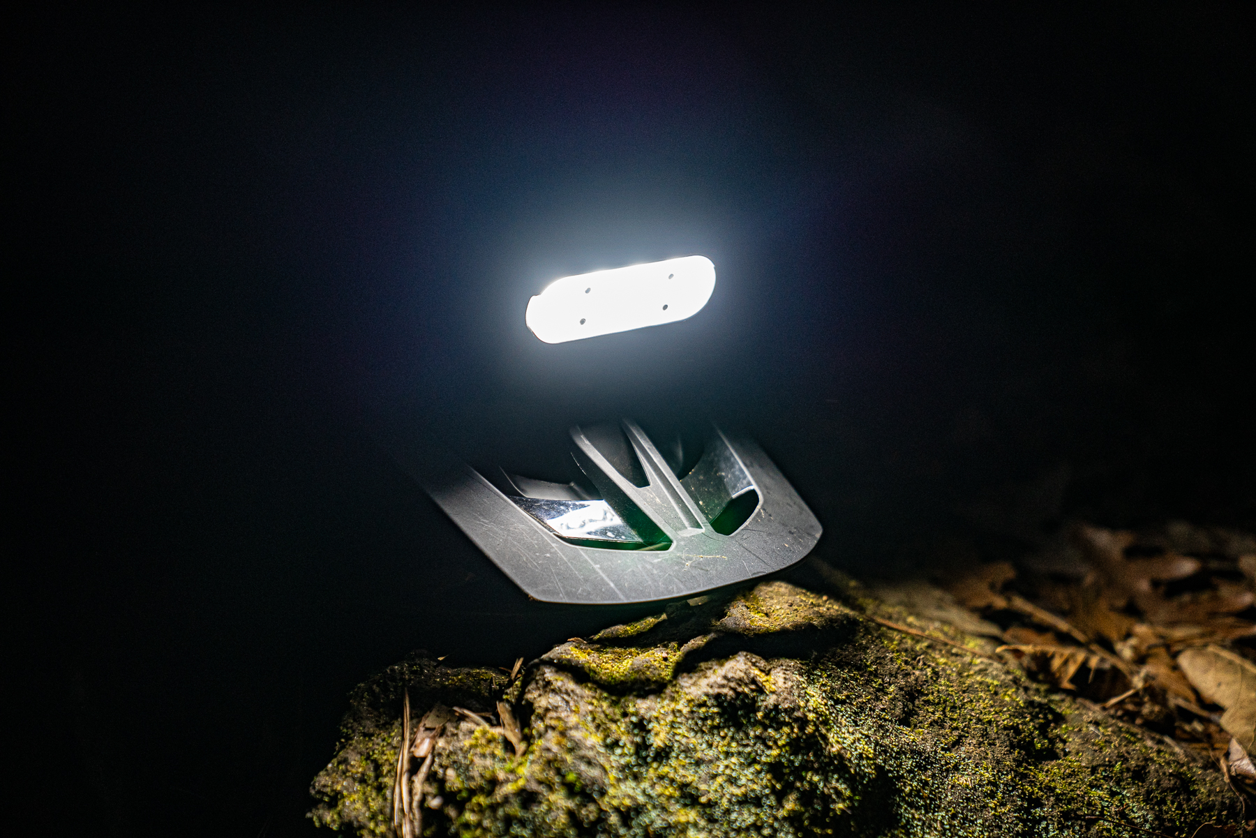 A focused beam of light illuminates a dark area, highlighting a rugged surface covered in moss and fallen leaves. The light source appears to be part of a larger object, featuring a sleek design. The surrounding environment is dimly lit, creating a contrast between the brightness of the light and the darkness around it.