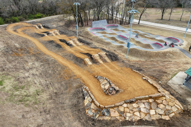 Aerial view of a dirt bike park featuring a winding dirt track with jumps on the left and a concrete pump track with several undulating mounds on the right. Several trees are in the background, and a small shelter is visible in the lower right corner. The park appears to be set in a natural landscape with a mix of grass and dirt terrain. Binkley mountain bike trail.