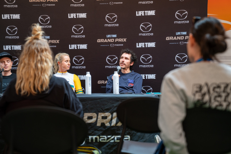 A panel discussion featuring three individuals seated at a table with branding from Mazda and Life Time. The participants include a man in a black cap (Lachlan Morton) and a man in a blue shirt speaking into a microphone, alongside a woman in a yellow and white outfit. In the foreground, the back of an audience member can be seen, focusing on the panelists. The backdrop displays promotional logos for Mazda and Life Time Grand Prix.
