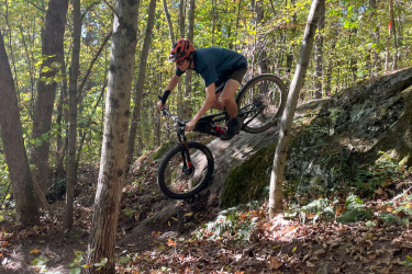 A mountain biker maneuvering down a rocky slope in a forest during autumn, surrounded by trees and fallen leaves, wearing a helmet and athletic gear. Maple Wind Farm mountain bike trail.