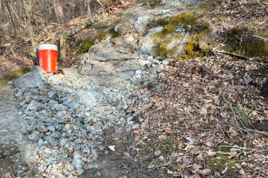 An area of a forest trail featuring a rocky embankment with a pile of gravel and a bright red bucket positioned beside it. The ground is covered with fallen leaves and patches of moss, surrounded by bare trees indicating early spring or late fall. Maple Wind Farm mountain bike trail.