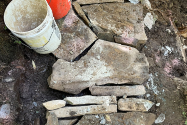 A view from above showing a stone pathway in progress, made from irregularly shaped rocks arranged on the ground. Two buckets, one orange and one white, sit nearby, and a person's feet can be seen at the bottom of the image, wearing muddy shoes. The surrounding area is partially shaded with dirt and greenery. Maple Wind Farm mountain bike trail.