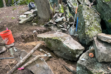 An excavation site in a wooded area, featuring large rocks and soil. A red bucket is positioned nearby, along with various tools including a shovel and a pickaxe, placed against a moss-covered rock. Pink flags are visible, marking specific locations in the digging area. The background shows trees and more rocky terrain, suggesting an active outdoor project. Maple Wind Farm mountain bike trail.