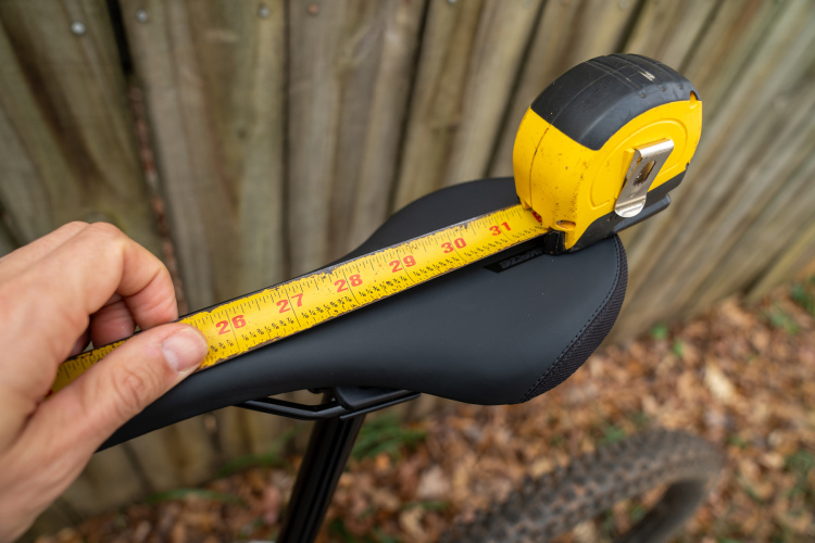 A close-up view of a bicycle seat being measured with a yellow tape measure. One hand is holding the tape measure against the seat, while the background shows a wooden fence and fallen leaves. The measurement indicates a length of approximately 27 inches.