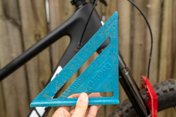 A hand holding a blue triangular scale ruler in front of a black bicycle frame against a wooden fence background.