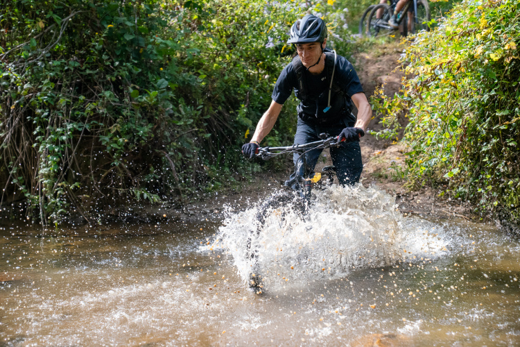 A mountain biker rides through a shallow stream, splashing water as he navigates the terrain. The surrounding area features lush greenery and sunlight filtering through the leaves. Another biker can be seen in the background.