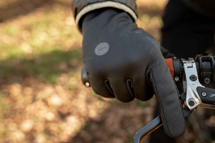 A close-up of a black-gloved hand resting on a bicycle handlebar, with the index finger pointing forward. The background features blurred patches of brown leaves and greenery, indicating an outdoor setting.