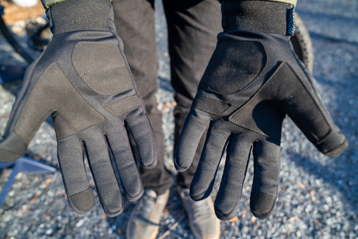 A person standing outdoors is holding up two black gloves, displaying them palm-up. The gloves have a textured design and are made of a flexible material, suitable for outdoor activities. The background features a gravel surface and a hint of equipment.