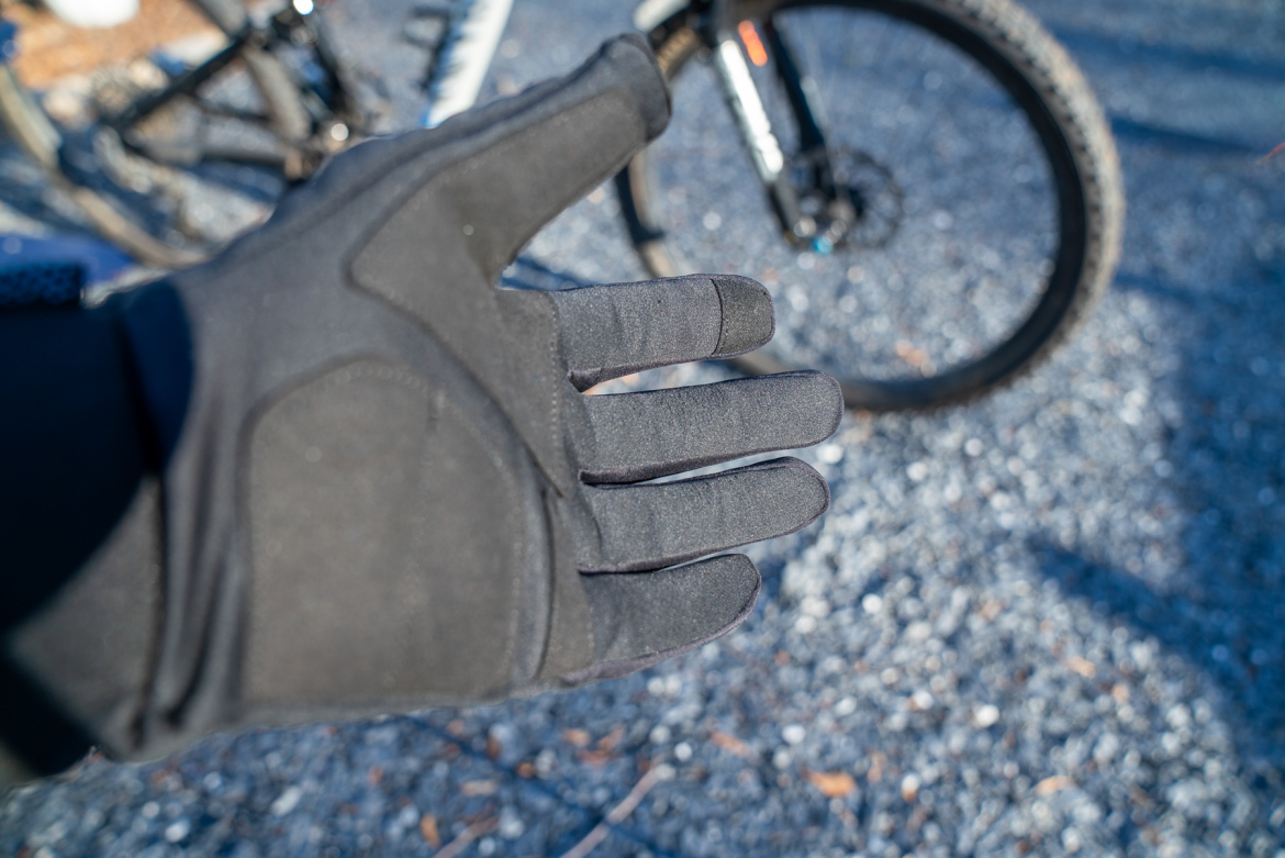 A close-up of a black glove held up, revealing the palm and fingers. In the background, a blurred black bicycle tire is partially visible on a gravel surface, suggesting an outdoor activity like cycling.