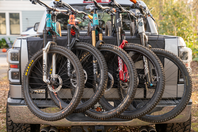 A collection of mountain bikes mounted on the back of a pickup truck, showcasing their handlebars and tires. The truck has a black protective cover on the tailgate, and the image captures the bikes in a natural outdoor setting.