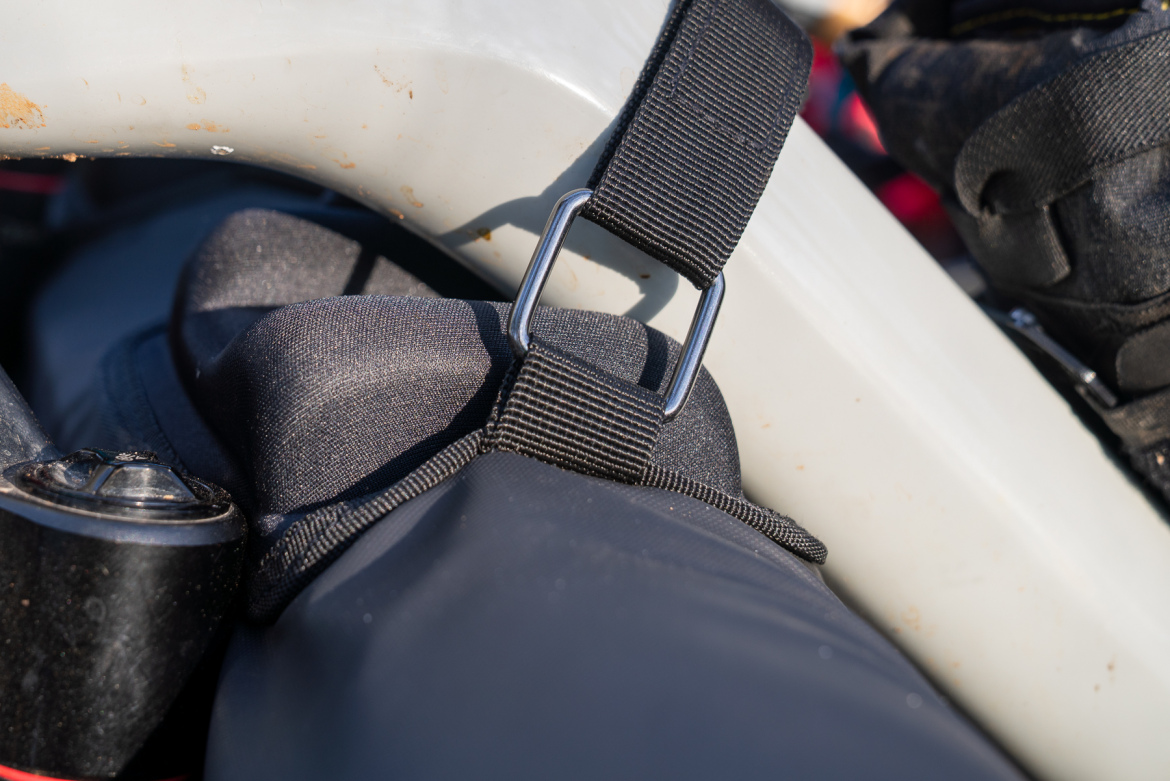Close-up image of a gray kayak frame showing a black strap with a metal buckle secured to a padded section. The background features parts of the kayak, highlighting the attachment point and gear setup.