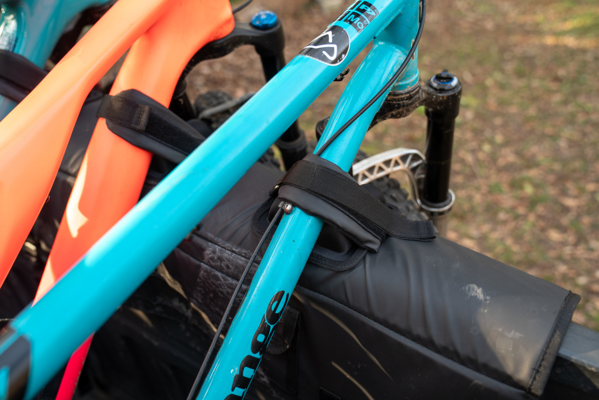 Close-up of bicycle components, featuring a turquoise bike frame secured with black straps, alongside an orange accessory. The background shows a blurred natural setting with leaves on the ground.