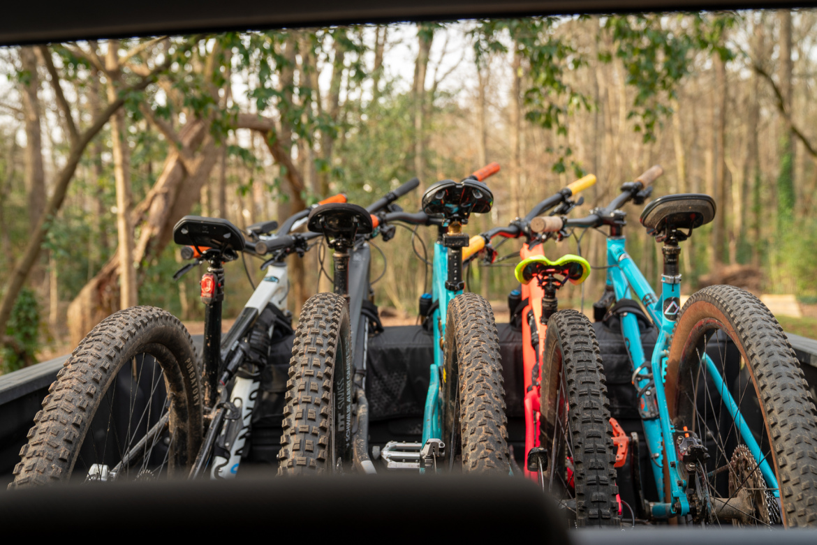 A view from inside a vehicle looking out at a row of mountain bikes loaded in the back, with their tires visible and the forest in the background.