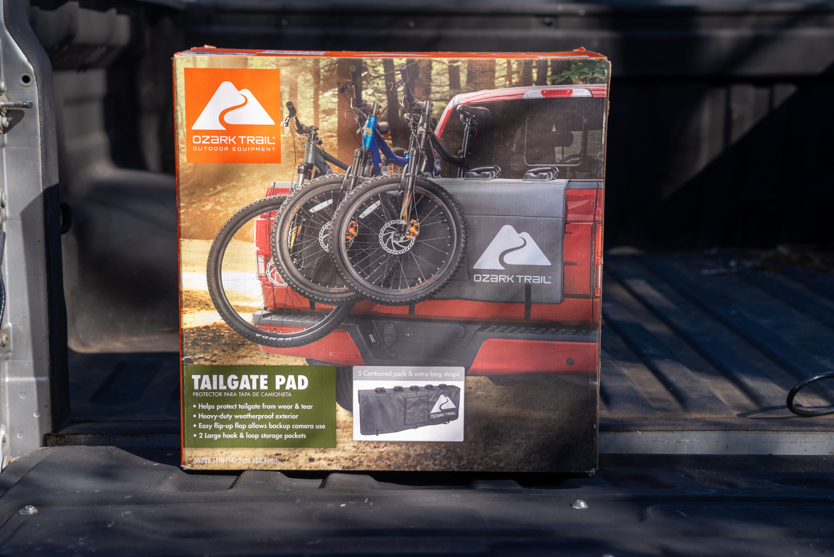 A box for an Ozark Trail Tailgate Pad, displayed in the back of a truck. The box features images of the tailgate pad with bicycles secured on it and highlights its protective features and waterproof exterior. The background shows a wooded area, emphasizing outdoor use.