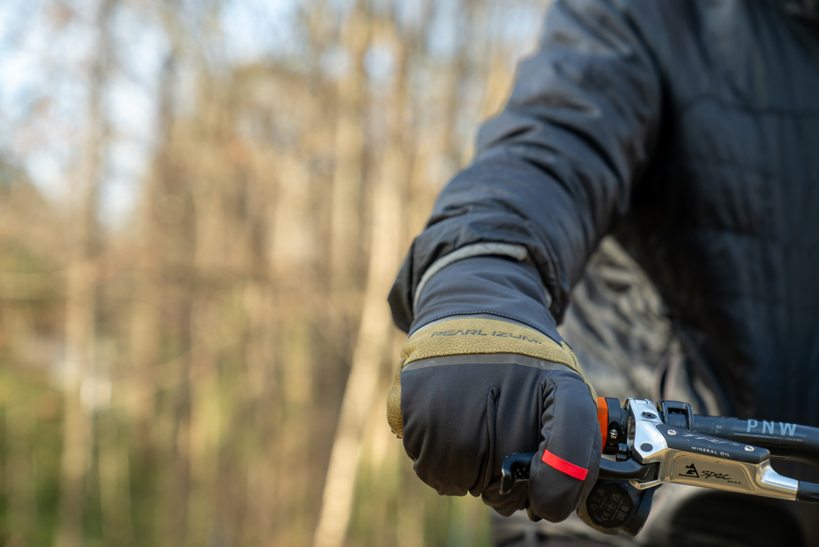 A close-up view of a hand wearing a black glove, gripping a bicycle handlebar. The glove has a brand logo visible, and a red strap is attached to the handlebar. In the background, there are blurred tree trunks, suggesting an outdoor setting.