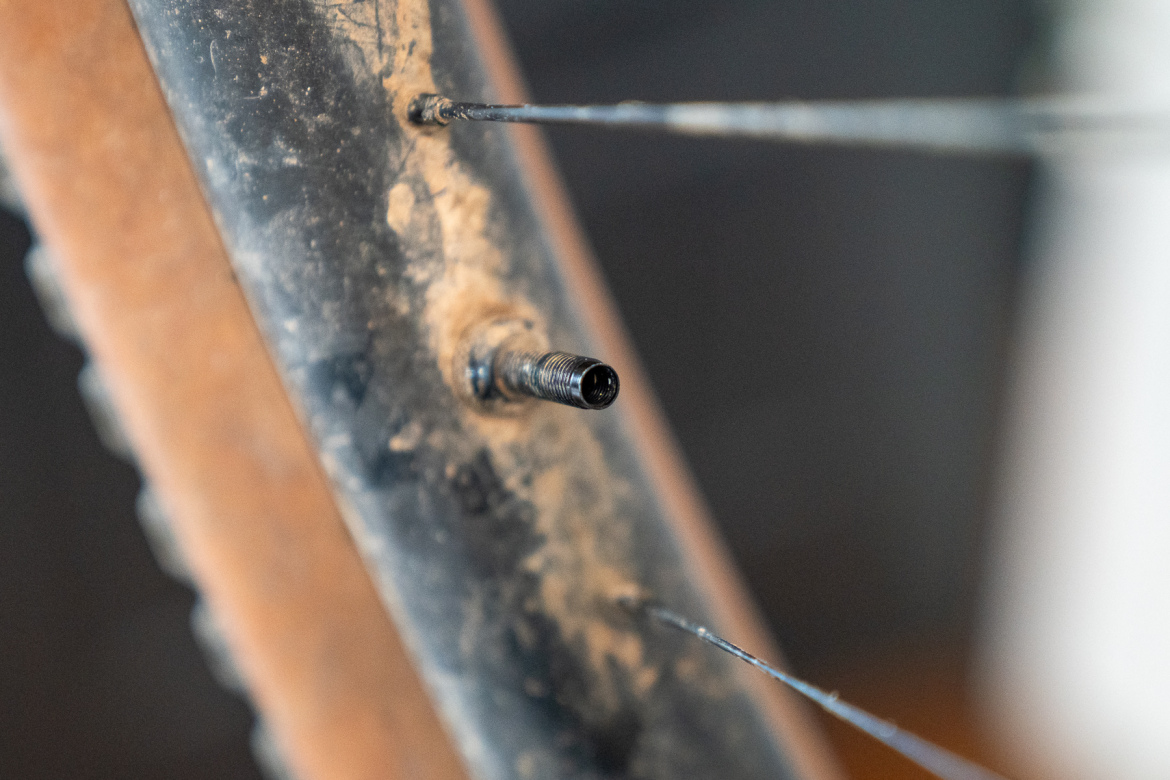 A close-up image of a bicycle tire valve on a dirty bike rim, with some wheel spokes visible in the background. The valve appears to be slightly worn and dusty, showing signs of use.