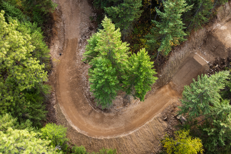 Aerial view of a winding dirt path surrounded by dense green trees, with a clearing visible alongside the curve of the trail.