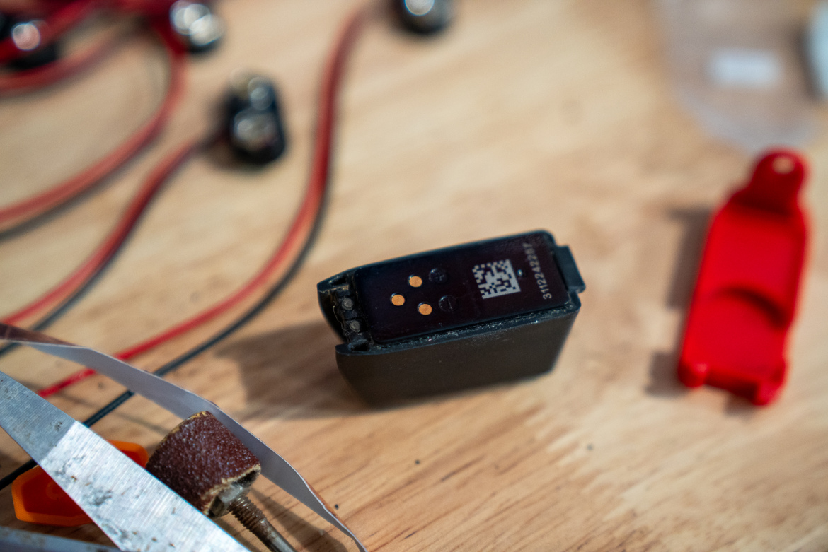 A close-up view of a small electronic component on a wooden surface, surrounded by wires and tools. The component features a black casing with a QR code and several small pins. Nearby, there is a red plastic cover and a piece of sandpaper, along with a pair of scissors and other wires.